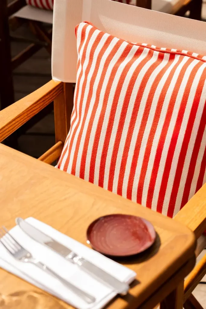 Chaise en bois avec coussin rayé rouge et blanc, couverts et assiette terracotta sur table de restaurant de plage