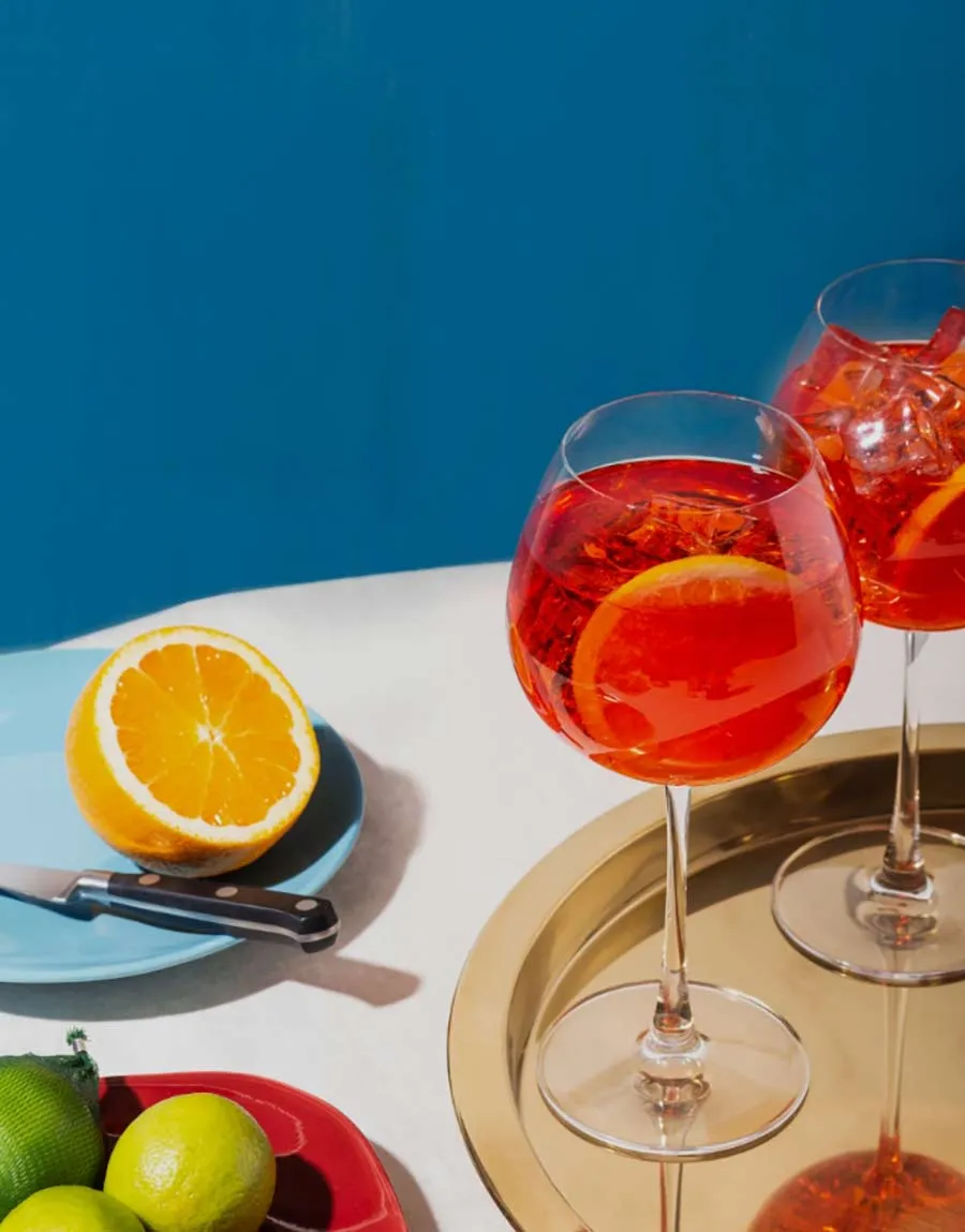 Deux verres d'Aperol Spritz sur plateau doré avec oranges et citrons verts, fond bleu azur