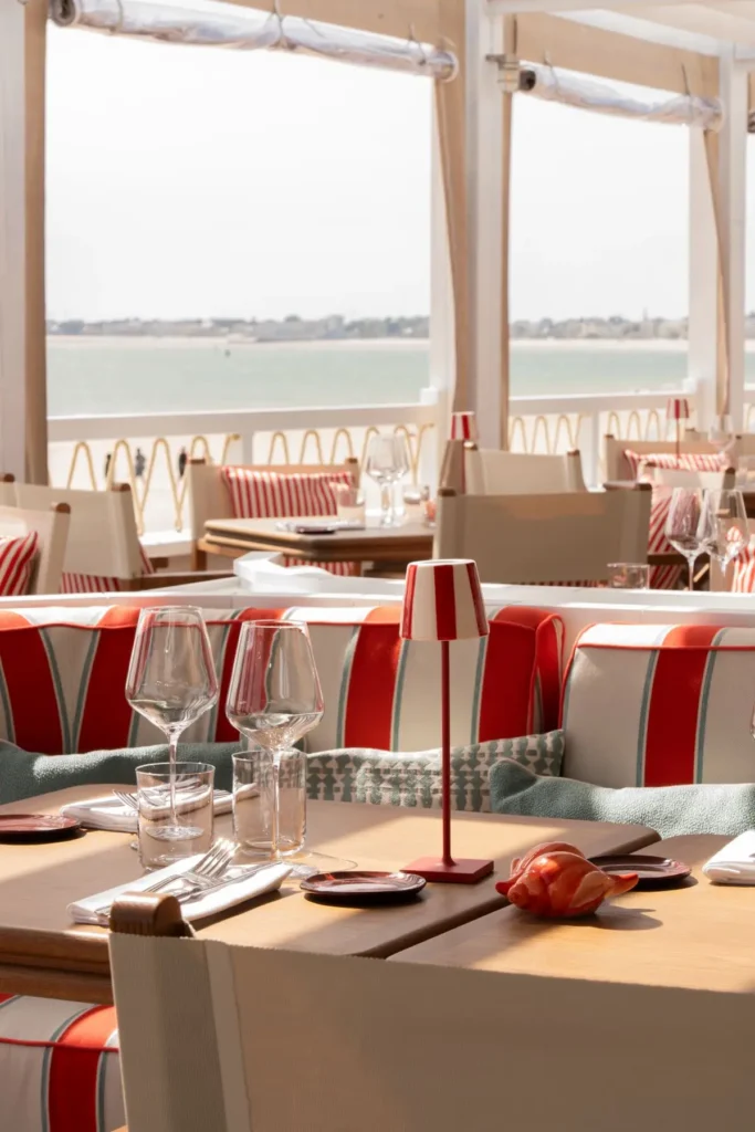 Tables dressées avec verres, lampes rayées rouges et banquettes colorées, vue sur la baie depuis les baies vitrées